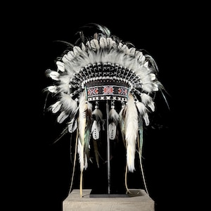 May include: A black and white headdress with feathers, beads, and a red and white beaded band. The headdress is on a black stand.