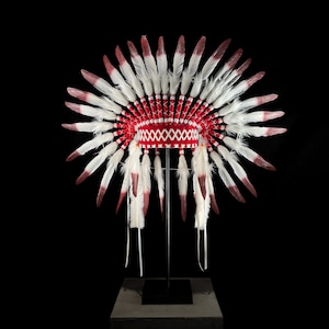 May include: A Native American headdress with a red and white design. The headdress features a red band with white stitching and is adorned with numerous white feathers with red tips. It is displayed on a black stand against a black background.