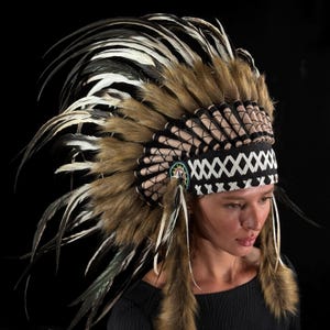 May include: A Native American headdress with brown, white, and black feathers. The headdress features a black band with white geometric designs and a beaded emblem. The feathers are a mix of brown, white, and black.