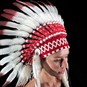 May include: A Native American headdress with white and red feathers. The headdress has a red band with white stitching and a white and red feather design. The feathers are white with red tips. The headdress is worn by a person.