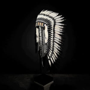 Op de afbeelding: Een traditionele Indiaanse hoofdtooi met witte en zwarte veren. De hoofdband heeft een zwart-wit kralenband met kleurrijke accenten. De veren zijn symmetrisch gerangschikt, wat een opvallend visueel effect creëert.