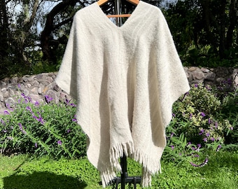 May include: A cream-colored knitted poncho with fringe hanging from the bottom edge. The poncho is hanging on a black stand with wheels.