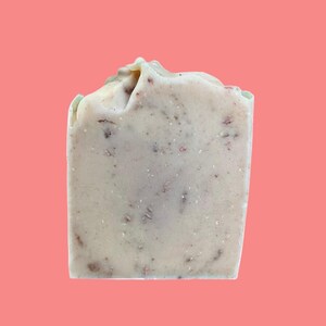 May include: A rectangular bar of handmade soap, pale beige in colour, with red and brown flecks. The top surface is textured and uneven. The background is a solid coral colour. This soap is likely for personal hygiene, and is suitable for washing.