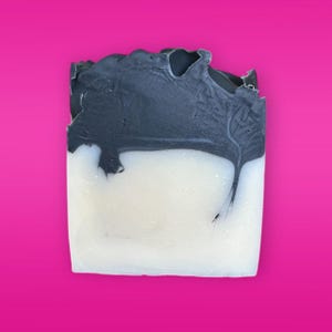 May include: A rectangular bar of soap with a two-tone design. The top section is a dark grey colour, while the bottom half is white. The soap has a textured, uneven surface, with the dark grey colour dripping down into the white.
