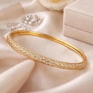 Peut inclure: Un bracelet jonc doré orné de diamants. Le bracelet est posé sur un tissu crème, avec d'autres bijoux en arrière-plan. La surface du bracelet est très réfléchissante et les diamants scintillent.