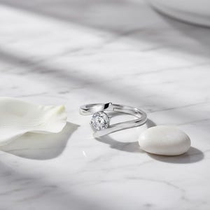 Peut inclure: Une bague en argent ornée d'une grosse pierre précieuse ronde et transparente. La bague présente un design ouvert unique avec un anneau fendu. Elle est présentée sur une surface en marbre blanc, à côté d'un pétale blanc et d'une pierre lisse blanche.