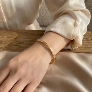 Peut inclure: Un bracelet doré avec un motif répété de petites pierres transparentes. Le bracelet est porté au poignet, avec une chemise de soie crème en arrière-plan. Le bracelet est un bijou simple et élégant.