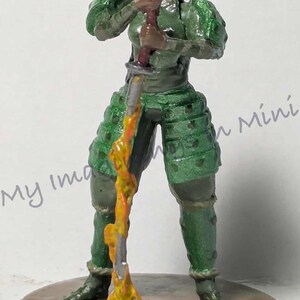 May include: A miniature figurine of a warrior in green armor, holding a sword with a yellow and orange flame effect. The figure stands on a brown base, with details like the armor's texture visible.