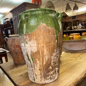 May include: A large, antique ceramic jar with a weathered appearance. The top is glazed in a vibrant green, with the glaze dripping down the sides onto the aged, brown clay. Two handles are visible on the sides. The jar is displayed on a wooden surface.