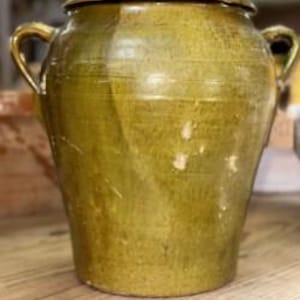 May include: A large, antique olive-green ceramic jar with a lid and two handles. The jar has a slightly textured surface with visible wear and tear, suggesting age. It sits on a wooden surface, and the background is blurred.