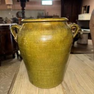 May include: A large, antique, olive-green ceramic jar with two handles. The jar has a wide, slightly flared rim and a textured surface. The jar is sitting on a wooden table. The background includes other antique items.