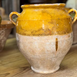May include: A large, antique ceramic pot with a wide, rounded body and two small handles. The top half is a vibrant yellow, while the bottom half is a lighter, beige color. The pot has a rustic, aged appearance, suggesting it is vintage.