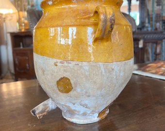 May include: Antique ceramic jug with a yellow glazed top and a cream-colored lower half. The jug features a handle and a spout, showcasing a rustic, aged appearance. This vintage piece is likely for decorative use.
