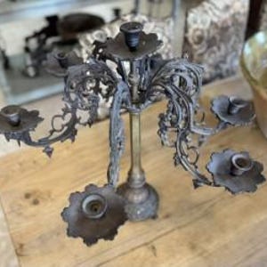 May include: An antique, ornate candelabra with six candle holders. The candelabra is made of metal with a tarnished, aged appearance. The candle holders are arranged in a multi-tiered design, with decorative floral accents. It sits on a wooden surface.
