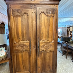 May include: A tall, two-door wooden armoire featuring a rich brown finish, arched top, and intricate carved floral rosette details on the panels. The cabinet includes vertical metal hardware along the door edges and stands on short, curved feet.