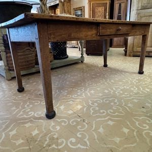May include: A rectangular wooden table with a natural brown finish, featuring a single drawer with a small round knob centered on the front apron. The table stands on four straight legs, each ending in a dark, rounded, ball-shaped foot base.