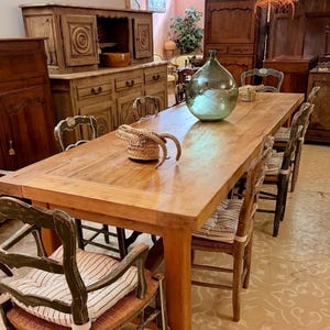 May include: A rectangular wooden dining table with a natural finish, surrounded by several chairs with green painted frames and woven seats. Each chair features a striped cushion. A large, clear glass demijohn bottle sits on the table surface.