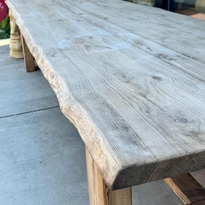 May include: A large, weathered wooden table with a natural edge. The table has a light gray color with visible wood grain and knots. The legs are made of a lighter-toned wood. This rustic table is suitable for dining or display.