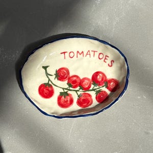 Op de afbeelding: Een handgemaakte keramische schaal met een witte basis en een blauwe rand. De schaal heeft een handgeschilderd ontwerp van rode tomaten op een wijnstok, met het woord "TOMATOES" in rode letters.