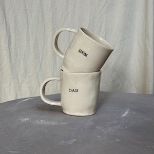 Puede incluir: Dos tazas de cerámica color crema apiladas una encima de la otra. La taza superior tiene la palabra "MOM" impresa, y la inferior "DAD". Ambas tazas tienen un asa curvada.