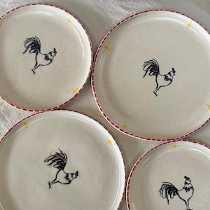 May include: Four off-white ceramic plates with a black rooster illustration in the center. Each plate has a red dotted border and yellow starburst accents. A charming addition to any kitchen.