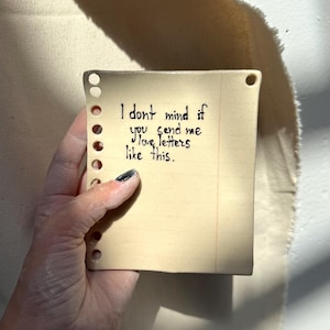 May include: A square, off-white ceramic piece resembling a notepad page. It has a lined paper design and holes along the left edge. The handwritten text reads, "I don't mind if you send me love letters like this."