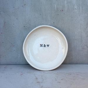 Peut inclure: Une assiette ronde en céramique blanc cassé avec un bord légèrement surélevé. L'assiette a une texture mouchetée et présente les lettres noires "M & W" centrées sur la surface. L'assiette est posée sur un fond gris.