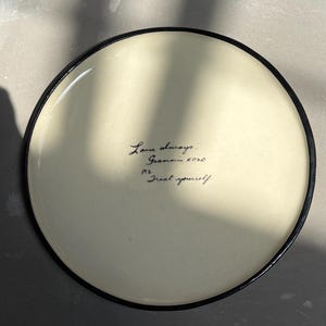 Könnte beinhalten: Ein runder, cremefarbener Keramiktarbener Keramiktfarbener Keramiktfarbener Keramikteller mit einem dünnen schwarzen Rand. In der Mitte befindet sich ein schwarzer, handschriftlicher Text: Love always, Grammie xoxo, P.S. Treat yourself. Die Oberfläche hat ein glattes, glänzendes Finish.