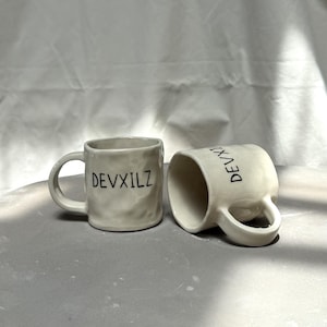 Könnte beinhalten: Zwei cremeweiße Keramikbecher mit einer strukturierten, unebenen Oberfläche und einem abgerundeten Henkel. Jeder Becher trägt den schwarzen Schriftzug DEVXILZ auf der Seite. Ein Becher steht aufrecht, während der andere daneben liegt.