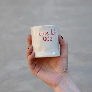 Könnte beinhalten: Ein kleiner, cremeweißer Keramikbecher mit einer leicht unregelmäßigen, handgeformten Gestalt. Der Text 'cute but OCD' ist in roter, handschriftlicher Schrift auf der Vorderseite zu sehen. Die Oberfläche des Bechers hat ein glänzendes, gesprenkeltes Finish.