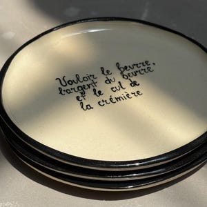 Könnte beinhalten: Ein Stapel von vier cremefarbenen Keramikplatten mit schwarzen Rändern. Jede Platte trägt ein handgeschriebenes Zitat in schwarzer Schrift. Die Teller haben eine glänzende Oberfläche und einen minimalistischen Stil.