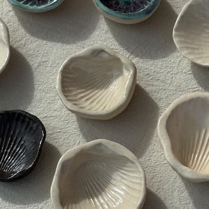 以下が含まれることがあります： さまざまな色と質感の、手作りの小さなセラミック皿のコレクション。貝殻の形をしたものにはリブのディテールがあり、丸いものには斑点または無地の仕上げが施されています。カラーパレットには、白、黒、ティールが含まれています。