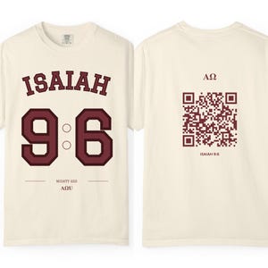 May include: An off-white t-shirt featuring a maroon graphic on the front with the text ISAIAH 9:6, MIGHTY GOD, and AΩ. The back of the shirt displays a maroon QR code with the text AΩ and ISAIAH 9:6 printed above and below the code.