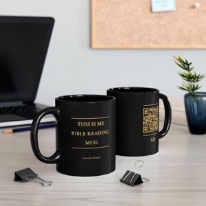 May include: Two black ceramic mugs featuring gold text and graphics. One mug displays the text 'THIS IS MY BIBLE READING MUG' and 'Gateway Designs'. The other mug features a gold QR code and the Greek letters Alpha and Omega in gold.