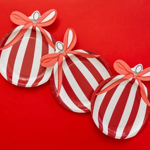 May include: Three red and white striped paper plates, each decorated with a bow and a silver-colored loop, arranged on a red background. The plates are designed to resemble Christmas ornaments.