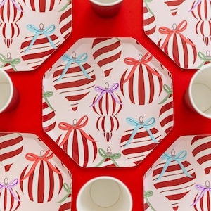 May include: Overhead shot of festive holiday plates and cups. The plates feature a cream background with red and white striped ornament designs, accented with colorful bows. Red cups are arranged around the plates on a bright red surface.