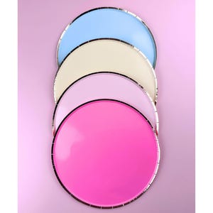 May include: Four colorful paper plates stacked on a pink background. The plates are light blue, cream, lavender, and hot pink. Each plate has a silver metallic rim. The plates are arranged in a diagonal line.