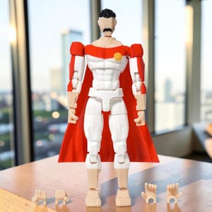 Könnte beinhalten: Eine Superhelden-Actionfigur mit rotem Umhang, weiß-rotem Anzug und austauschbaren Händen. Die Figur hat einen schwarzen Schnurrbart und Haare sowie ein gelbes Emblem auf der Brust. Die Figur steht auf einer Holzoberfläche.