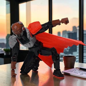 Könnte beinhalten: Detaillierte Figur eines futuristischen Kriegers in dynamischer Pose. Die Figur ist überwiegend schwarz mit roten und grauen Akzenten, einschließlich eines roten Umhangs. Der Krieger kniet auf einer Holzoberfläche.