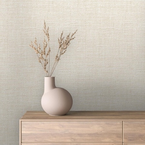 May include: A beige ceramic vase with a rounded body and a narrow neck holds dried wheat stalks. The vase sits on a light wood cabinet against a textured, off-white wall.