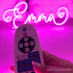 May include: A neon sign in the shape of the name "Emma" in a cursive font, illuminated in bright pink. A white remote control is in the foreground, with buttons for speed, light, and mode control.