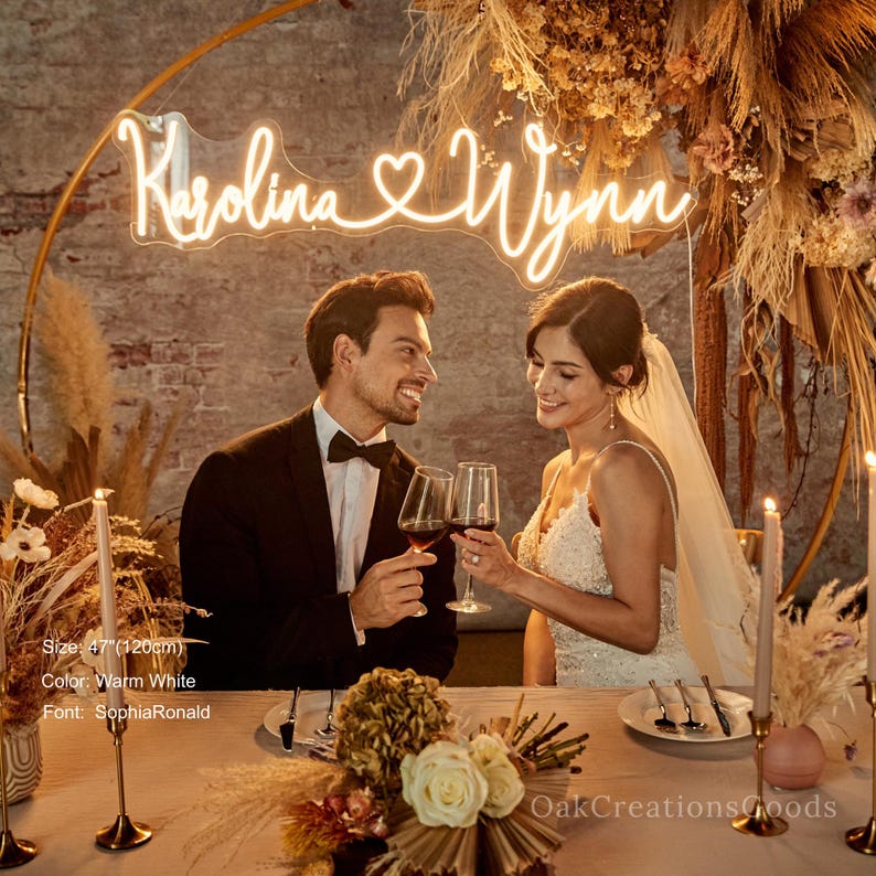 May include: A neon sign with the names "Karolina & Wynn" in warm white, 47 inches (120cm) wide, hangs above a couple toasting with wine glasses. The couple is seated at a table with floral arrangements and candles. The font is SophiaRonald.