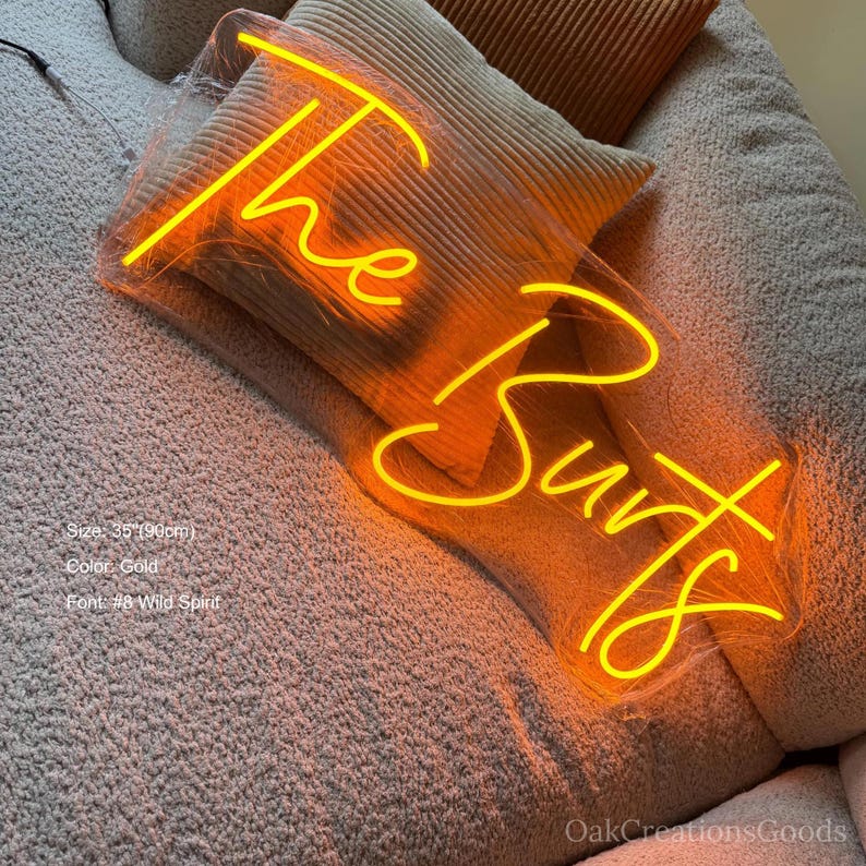 May include: A gold neon sign in the shape of an arrow, with the words "The Burts" in a cursive font. The sign is 35 inches (90cm) long and is placed on a beige surface with pillows.