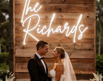 Puede incluir: Un letrero de neón blanco con la inscripción "The Richards" en escritura cursiva está montado en una pared de madera. Una pareja se encuentra frente al letrero, la mujer con un vestido de novia blanco y velo, el hombre con un esmoquin negro.