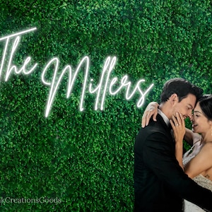 May include: A couple poses for a wedding photo in front of a green wall with a white neon sign that reads "The Millers".