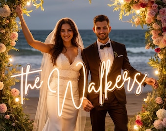 Puede incluir: Una pareja posa bajo un arco floral en una playa, sosteniendo un letrero de neón que dice "The Walkers". La mujer lleva un vestido de novia blanco y velo, el hombre un traje negro y pajarita. El arco está adornado con flores rosas y luces.