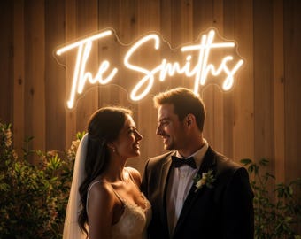 Puede incluir: Una pareja se mira frente a un letrero de neón que dice "The Smiths". El letrero está iluminado en un color blanco cálido, sobre un fondo de madera. La novia lleva un vestido de novia blanco y un velo, mientras que el novio viste un esmoquin negro.