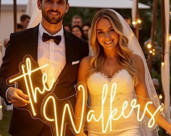 Puede incluir: Una pareja sonríe, sosteniendo un letrero de neón que dice "the Walkers" en una fuente cursiva. El letrero brilla con una luz amarilla cálida. El hombre viste un esmoquin negro con pajarita, y la mujer un vestido de novia blanco y velo.