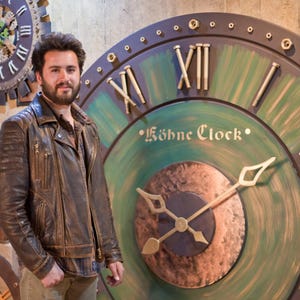 May include: A large, decorative clock with a green and bronze face, featuring Roman numerals and the text "Köhe Clock." The clock has a dark brown border and gold-coloured accents. The clock hands are white. The clock is mounted on a wall.