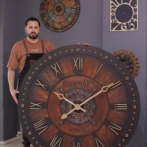 Puede incluir: Reloj de pared de madera grande y redondo con números romanos y el texto "BOHEMART BITLIS". El reloj tiene un anillo exterior marrón oscuro con detalles y manecillas doradas. Otros relojes son visibles en el fondo.